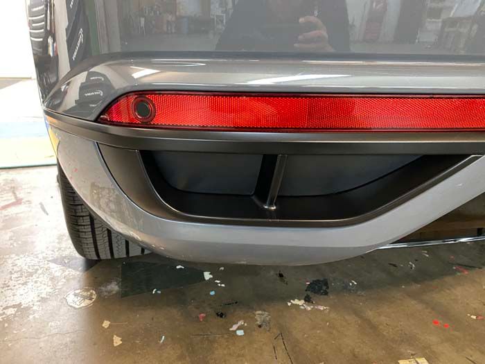Rear car bumper detail with a red reflector, gray paint, and a black trim.