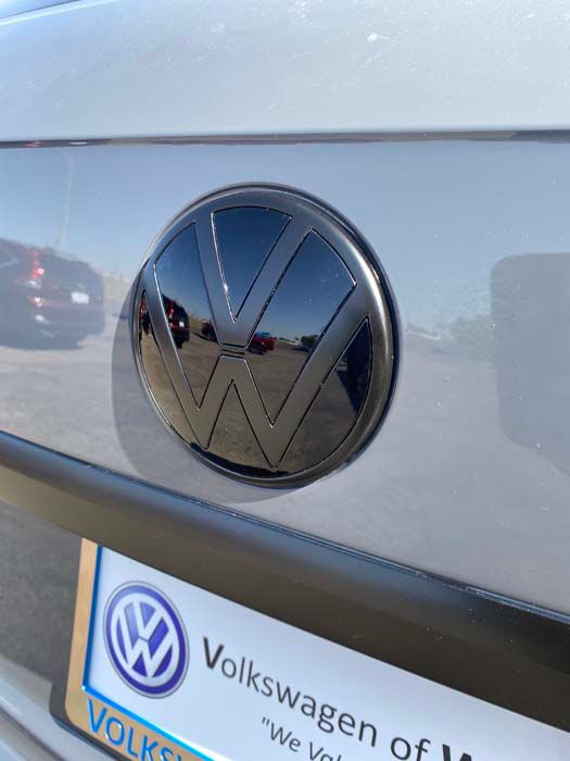 Black Volkswagen emblem on the back of a car, reflecting the environment, above a license plate.