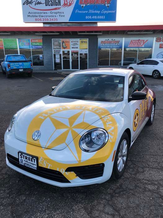 White Volkswagen Beetle with large yellow compass-like logo parked in front of a storefront.