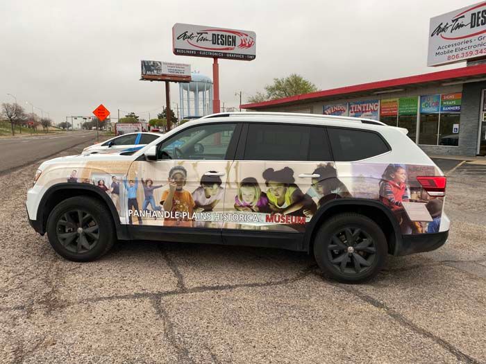 White SUV with photo wrap, black wheels, parked in front of a sign and building.