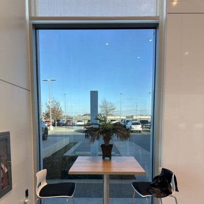 Interior view through a large window: table with plant, chairs. Outside, parking lot, blue sky.