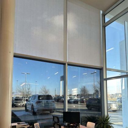 Large windows with white shades in a showroom; vehicles and blue sky reflected.