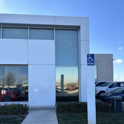 Exterior of a white building with large windows, a handicap parking sign, and cars parked outside on a sunny day.