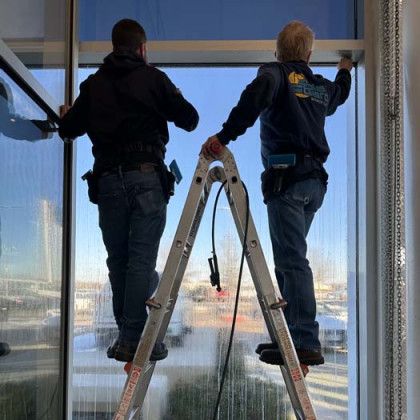 Two men installing window film on an office building window, using a ladder. Bright blue sky visible.