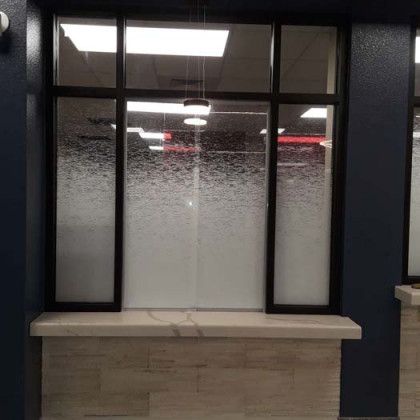 Window with frosted glass, set in a dark frame, above a stone counter. Inside, office lights and ceiling.