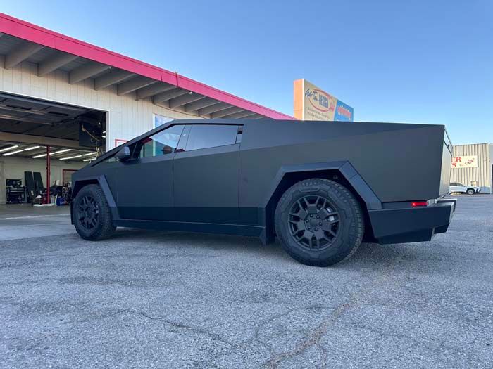 Tesla Cybertruck, matte black, parked outside a repair shop on a sunny day.