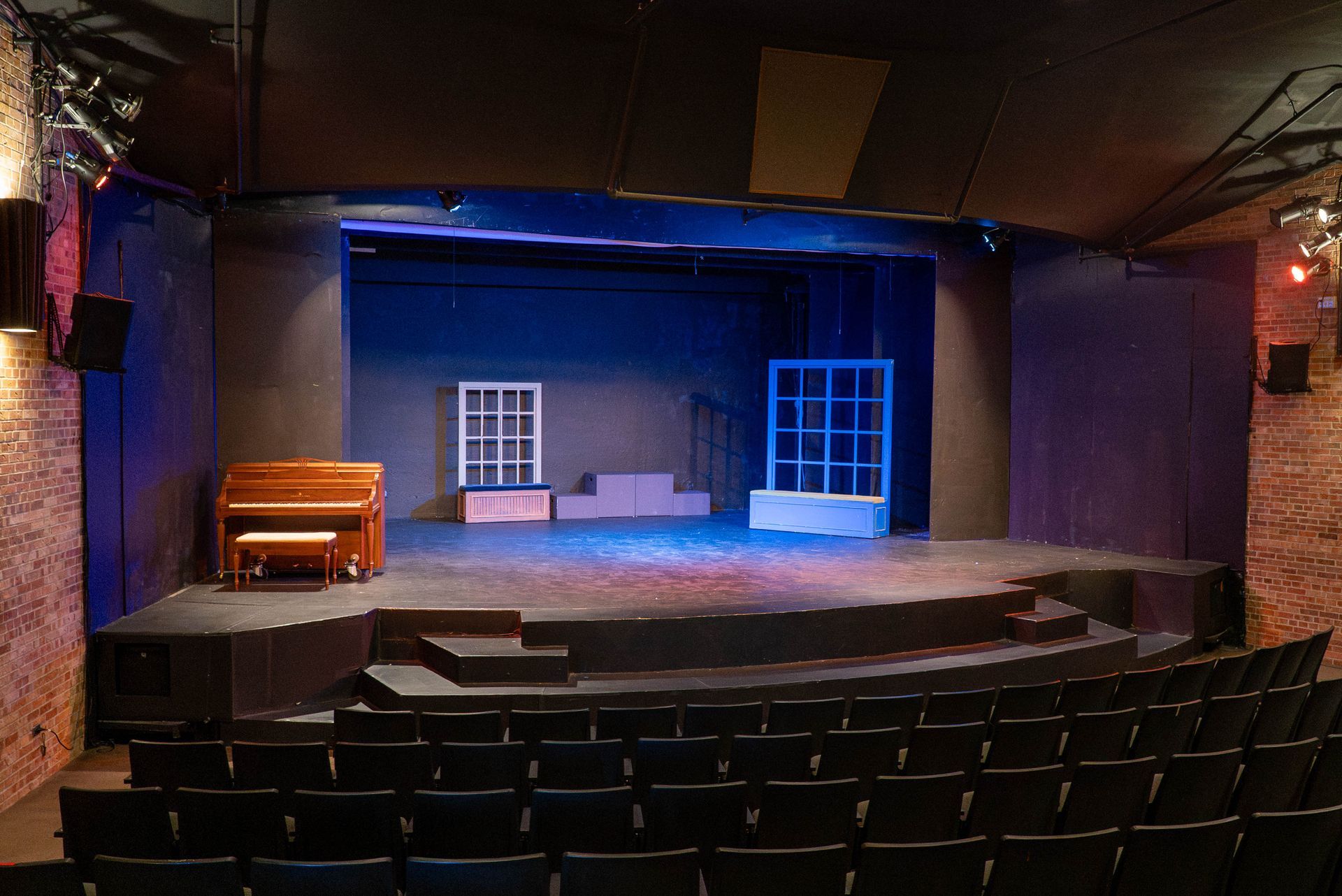 Empty theater stage with blue-lit set, piano at left, and black audience seats in foreground