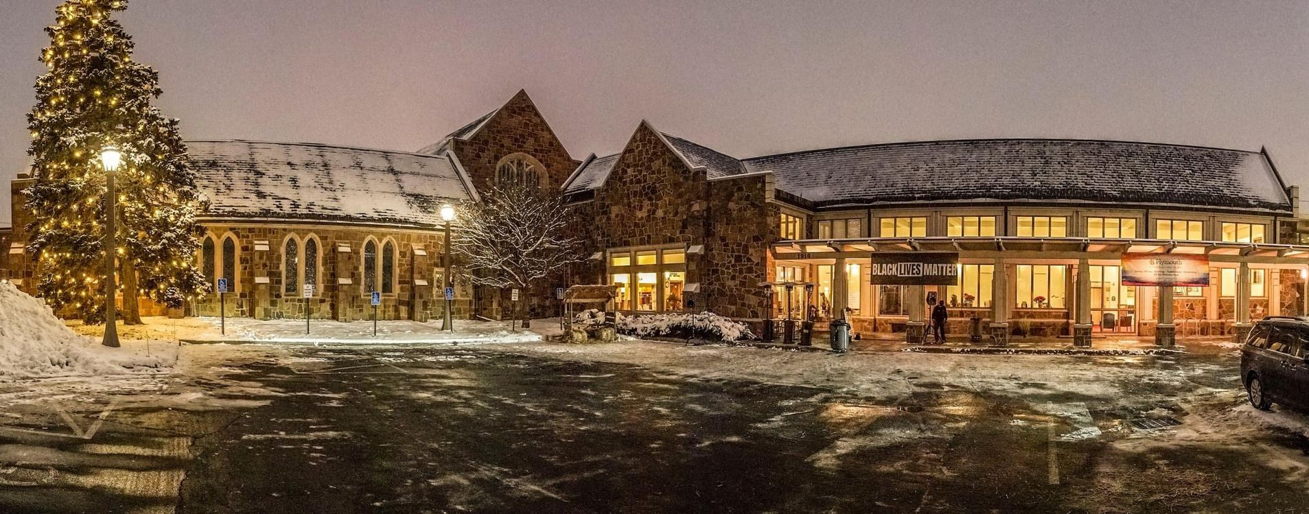 Snowy nighttime exterior of a lit building with decorated Christmas trees and warm windows