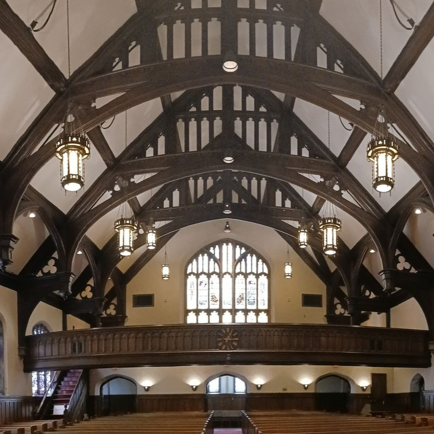 Church sanctuary with people seated in pews, facing a wood-paneled stage under arched beams.