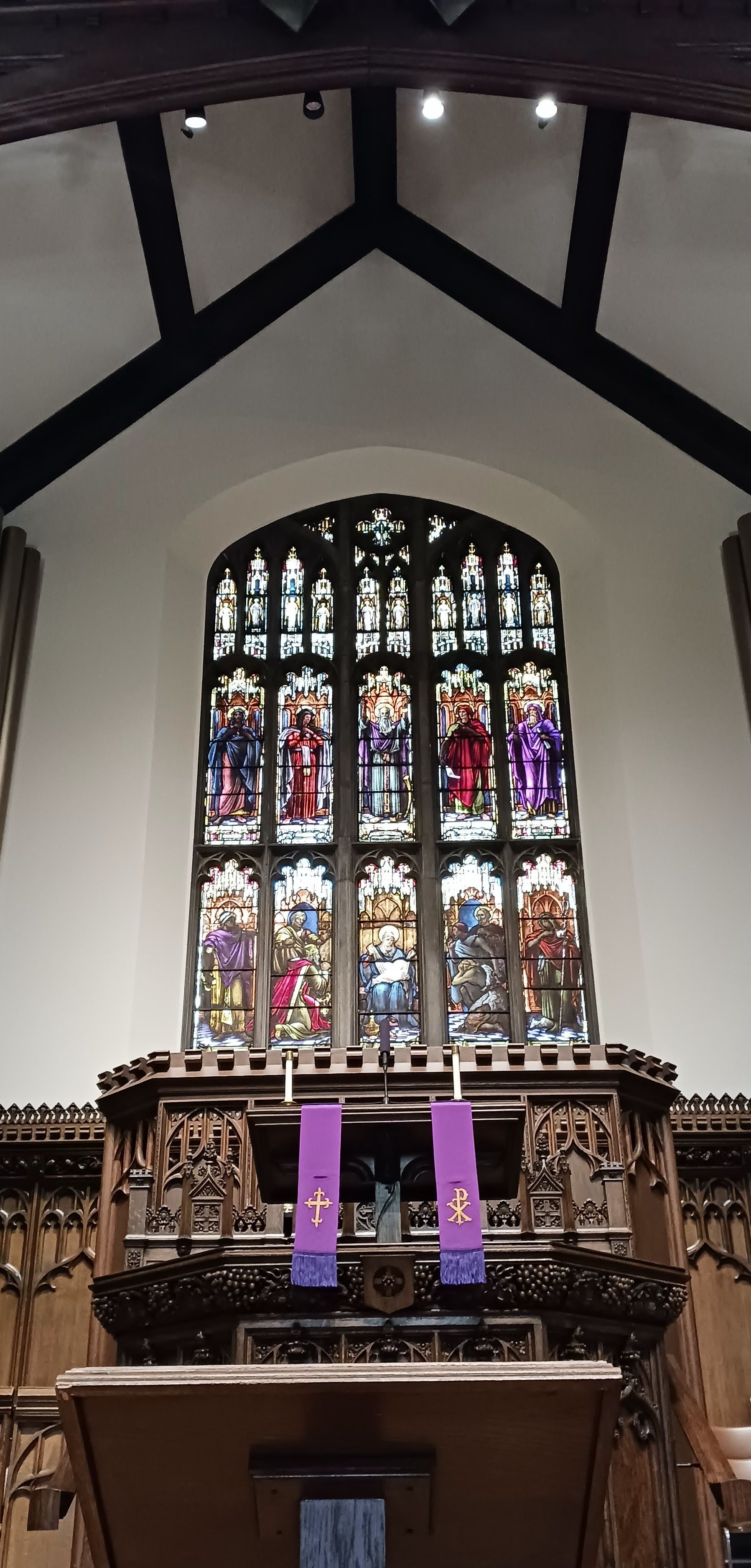 Church altar beneath arched stained-glass window, with purple banners and dark wooden beams above.