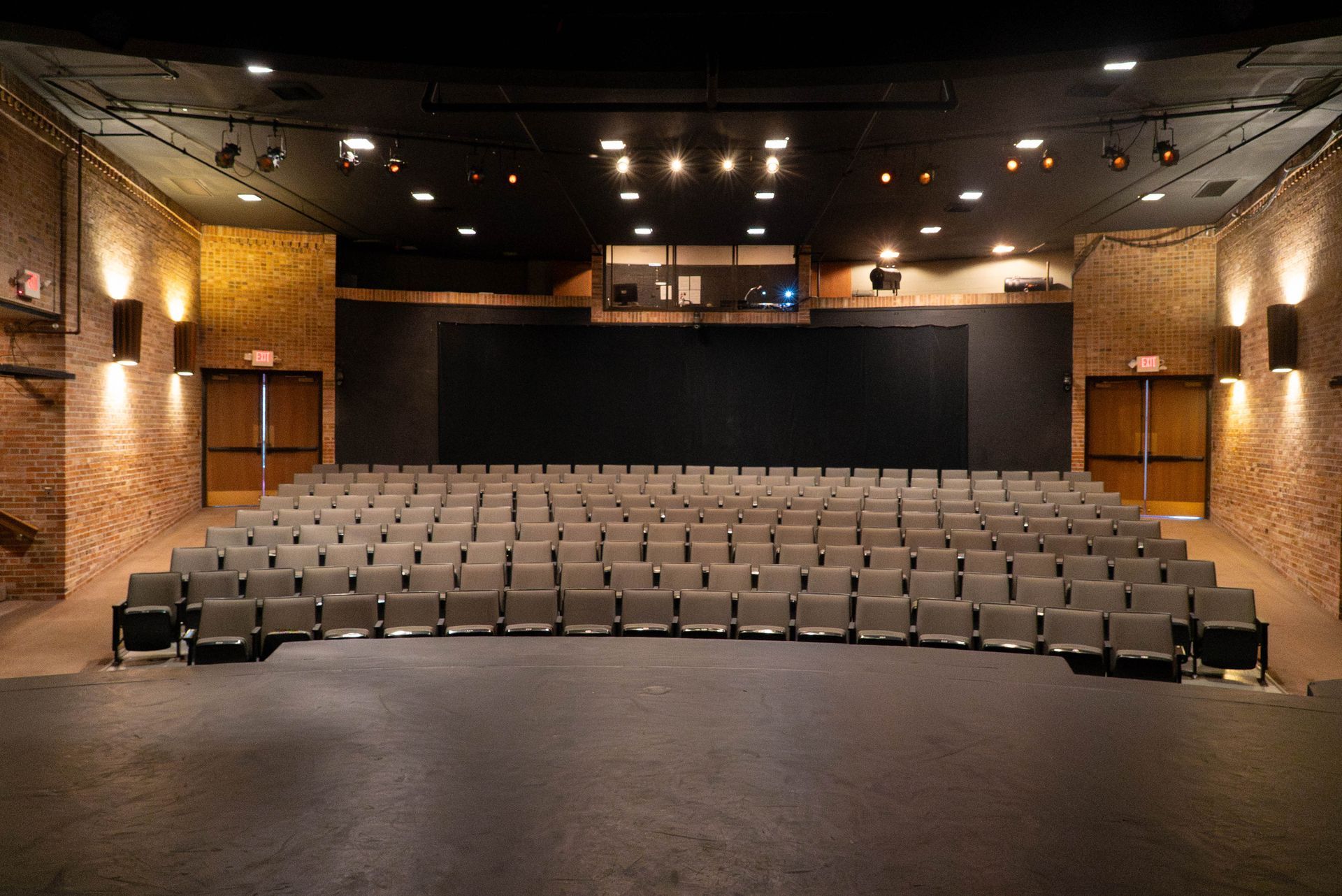Empty small theater with rows of gray seats facing a black stage and warm brick walls