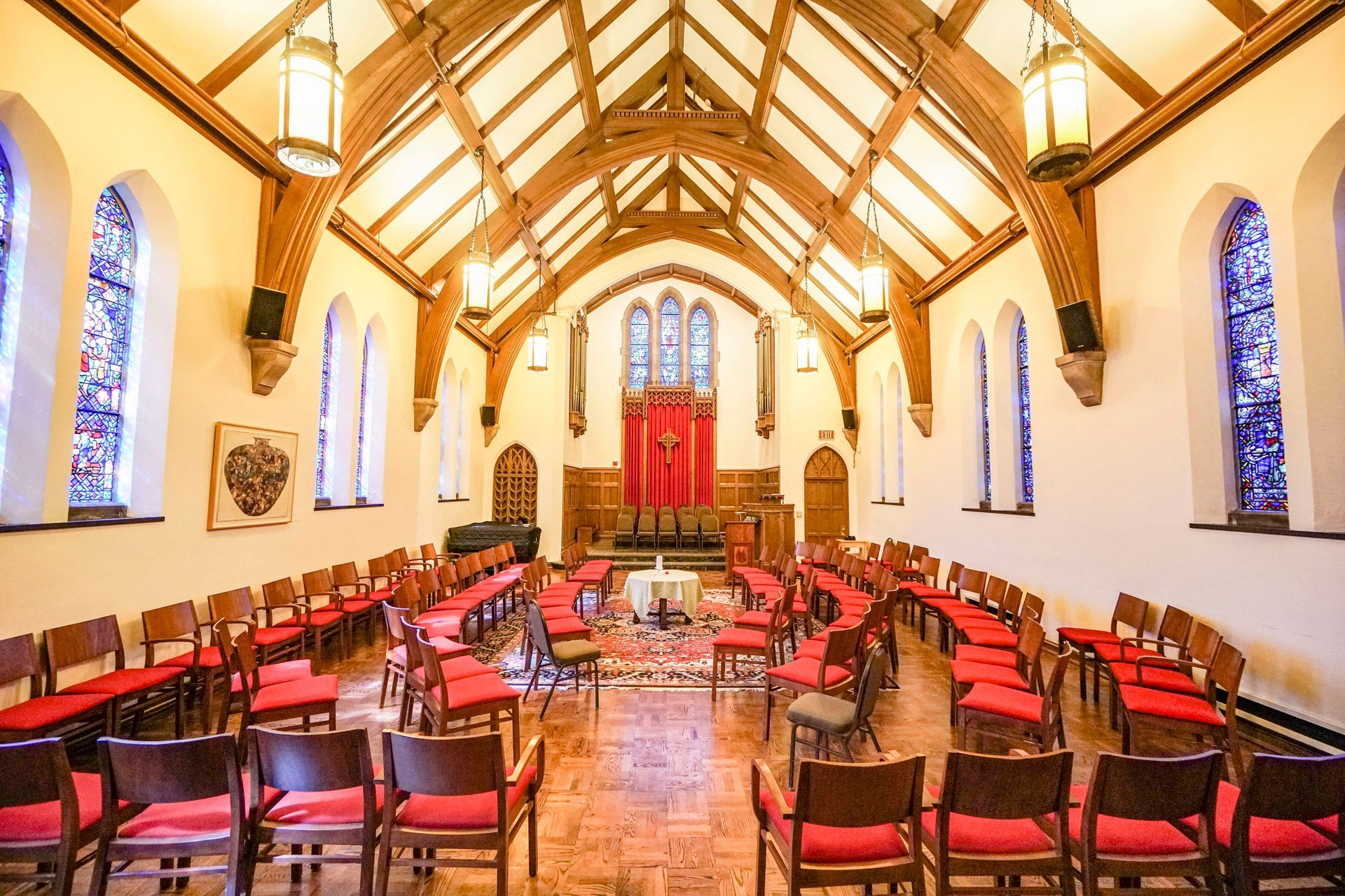 Bright church interior with red chairs arranged in rows and stained-glass windows on either side.