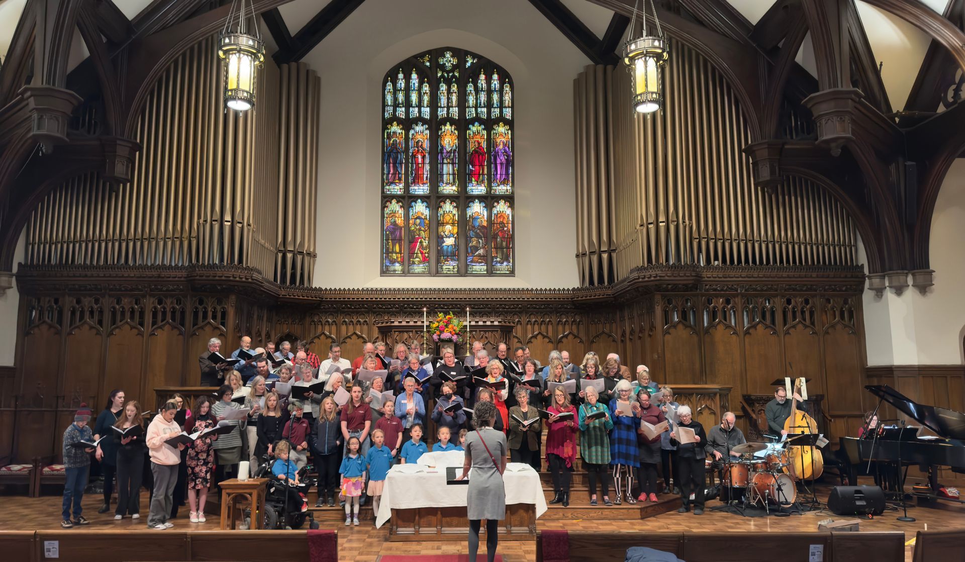 Choir and orchestra performing on a church stage beneath a large stained-glass window