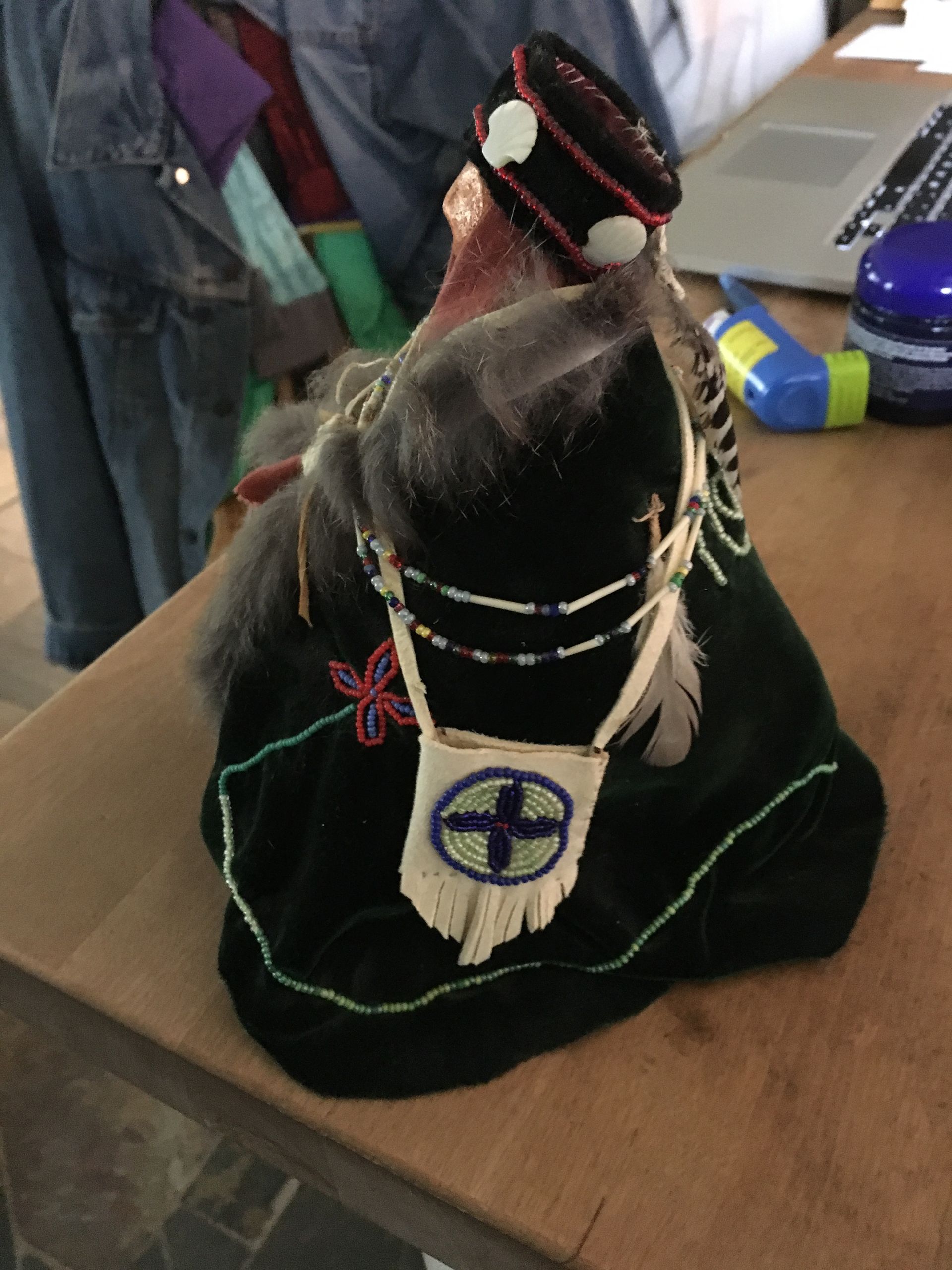 Decorated black cloth figurine with white feathers and beaded necklace on a tabletop