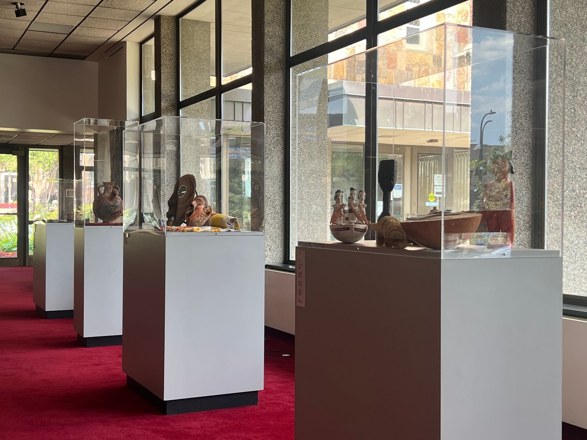 Gallery display with small sculptures on white pedestals in a bright room with large windows and red carpet