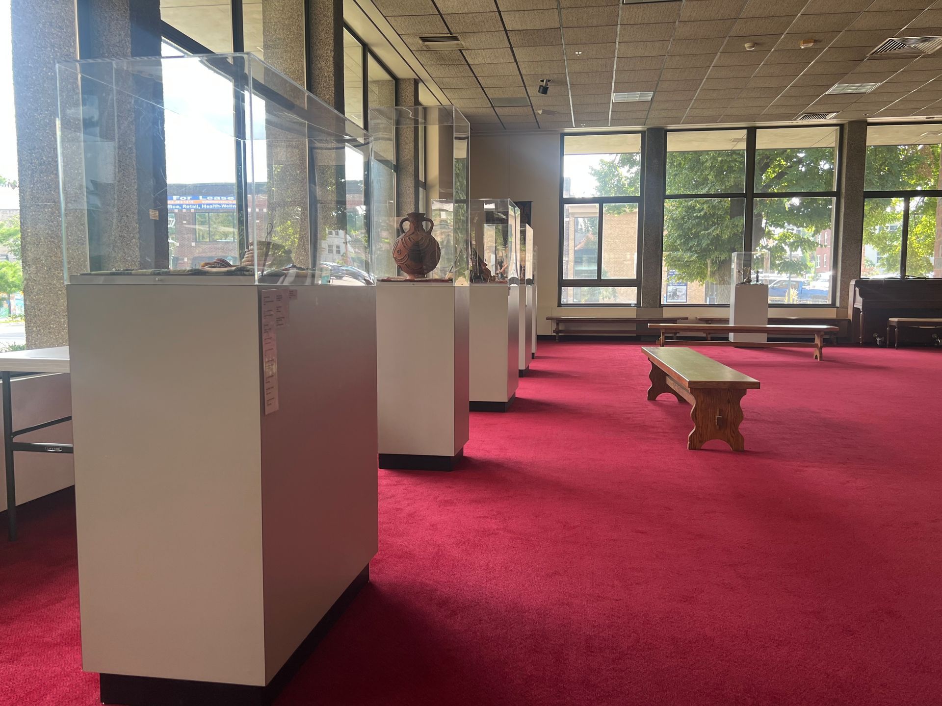 Bright lobby with red carpet, white display pedestals, and a wooden bench by large windows.