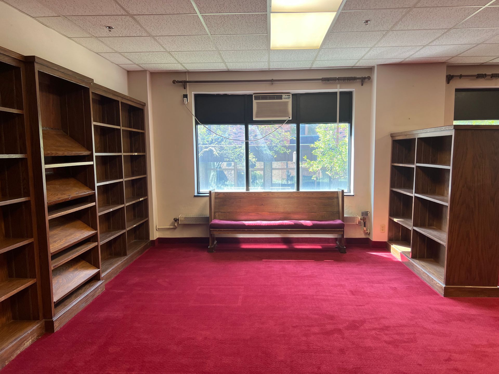 Empty room with red carpet, wooden shelves, and a bench by a window.