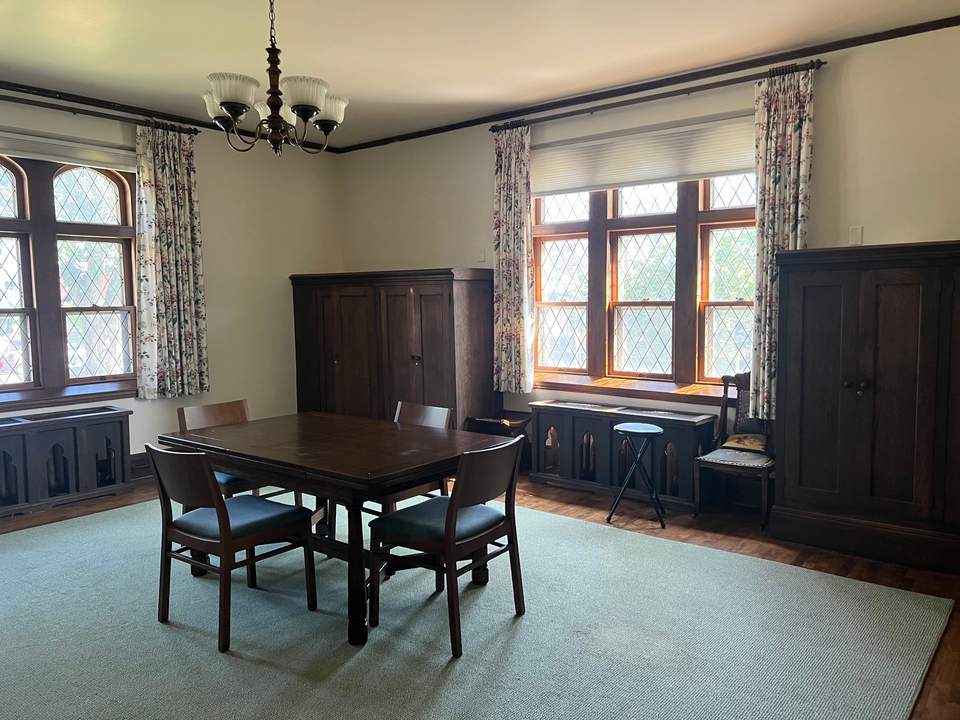 Empty dining room with wooden table, chairs, large windows, dark cabinets, and green carpet.