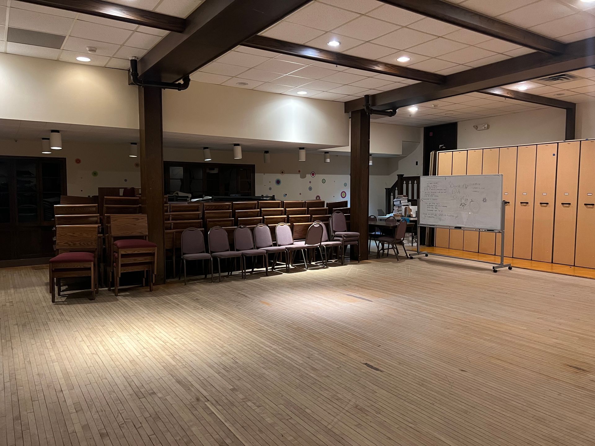 Empty multipurpose room with chairs, tables, whiteboard, and lockers under warm ceiling lights.