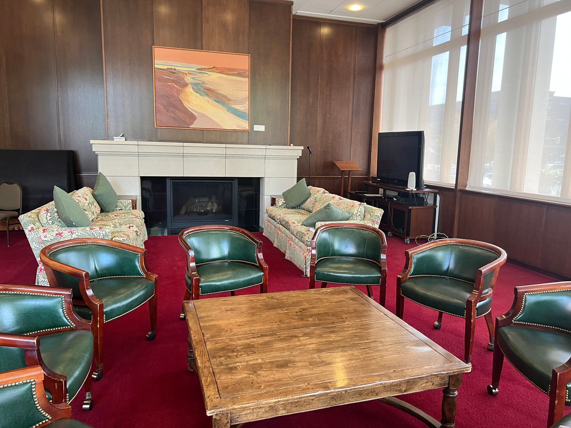 Living room with red carpet, green leather chairs, wooden table, and a fireplace under a landscape painting