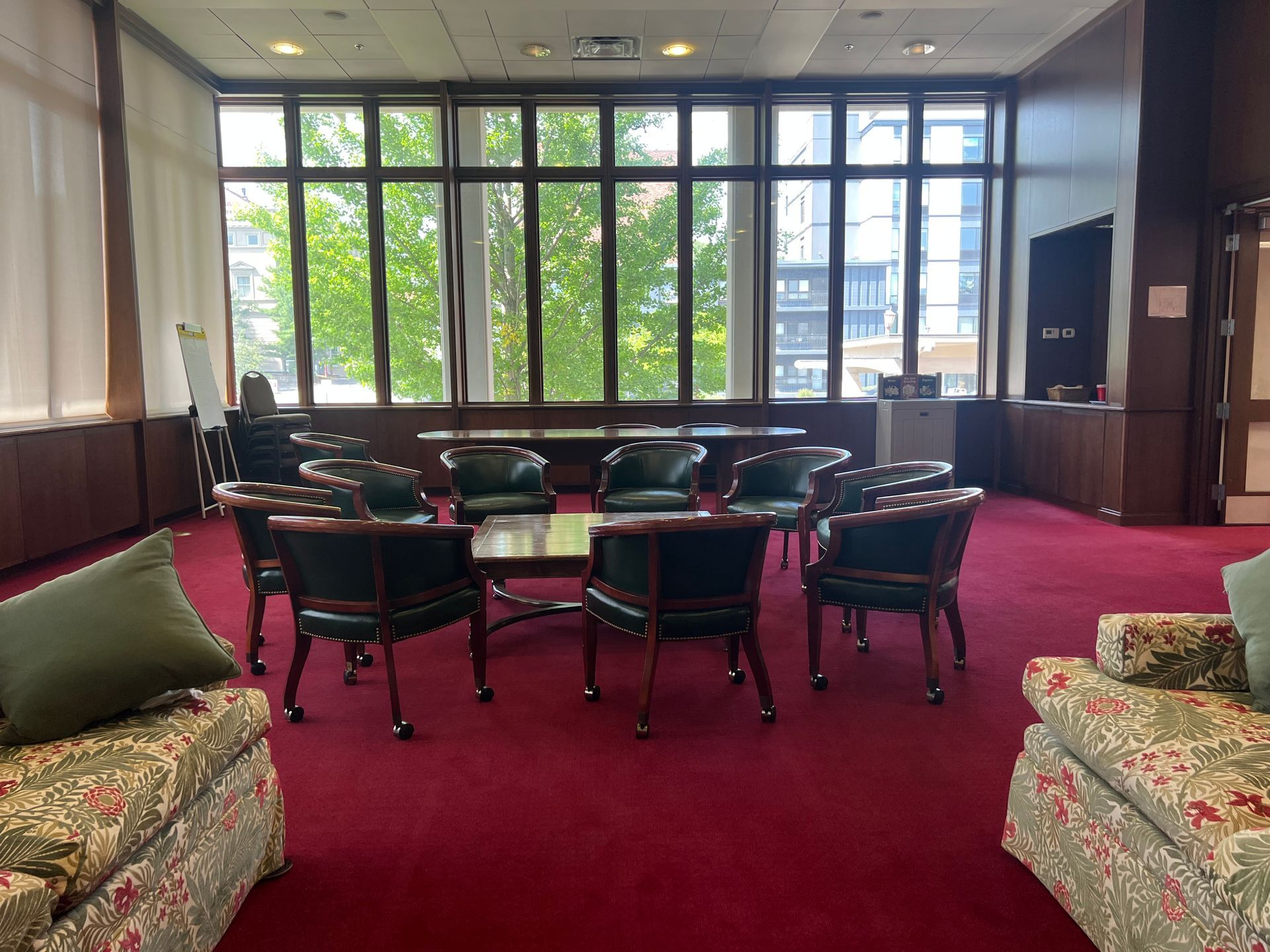 Lounge with red carpet, floral sofas, round tables, and large windows looking onto trees and buildings