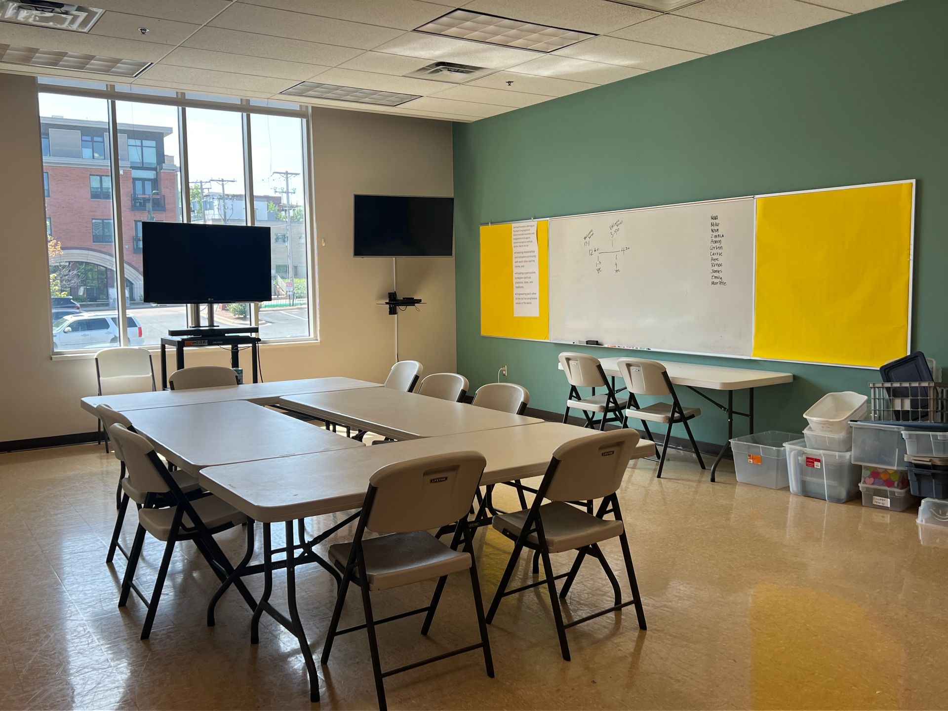 Bright classroom with tables, folding chairs, whiteboard, and yellow bulletin boards near large windows