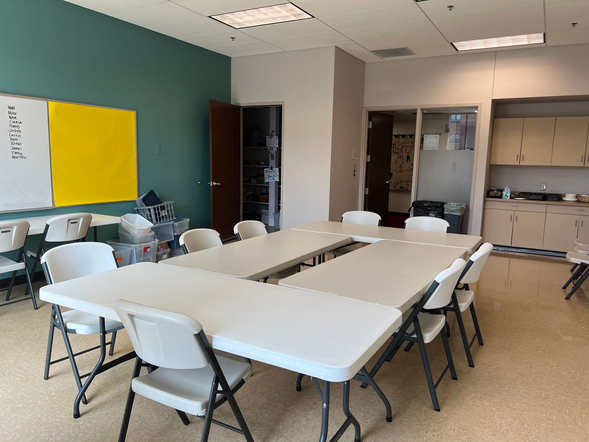 Empty classroom with white tables, folding chairs, teal wall, and a kitchen area in the back