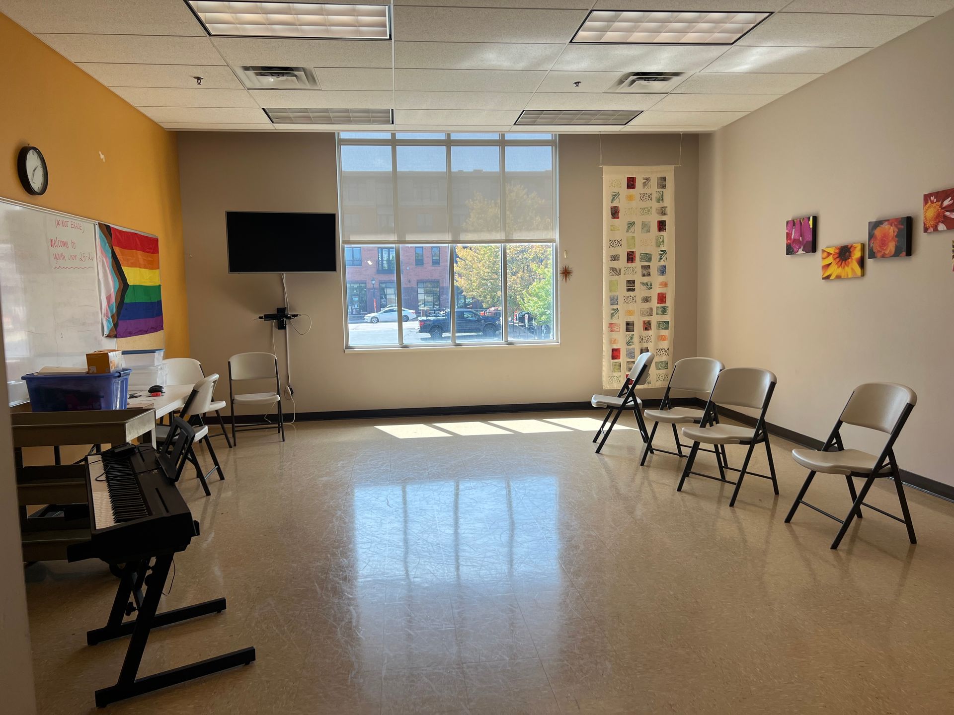 Empty classroom with chairs along the wall, a rainbow flag, and a large window letting in daylight.