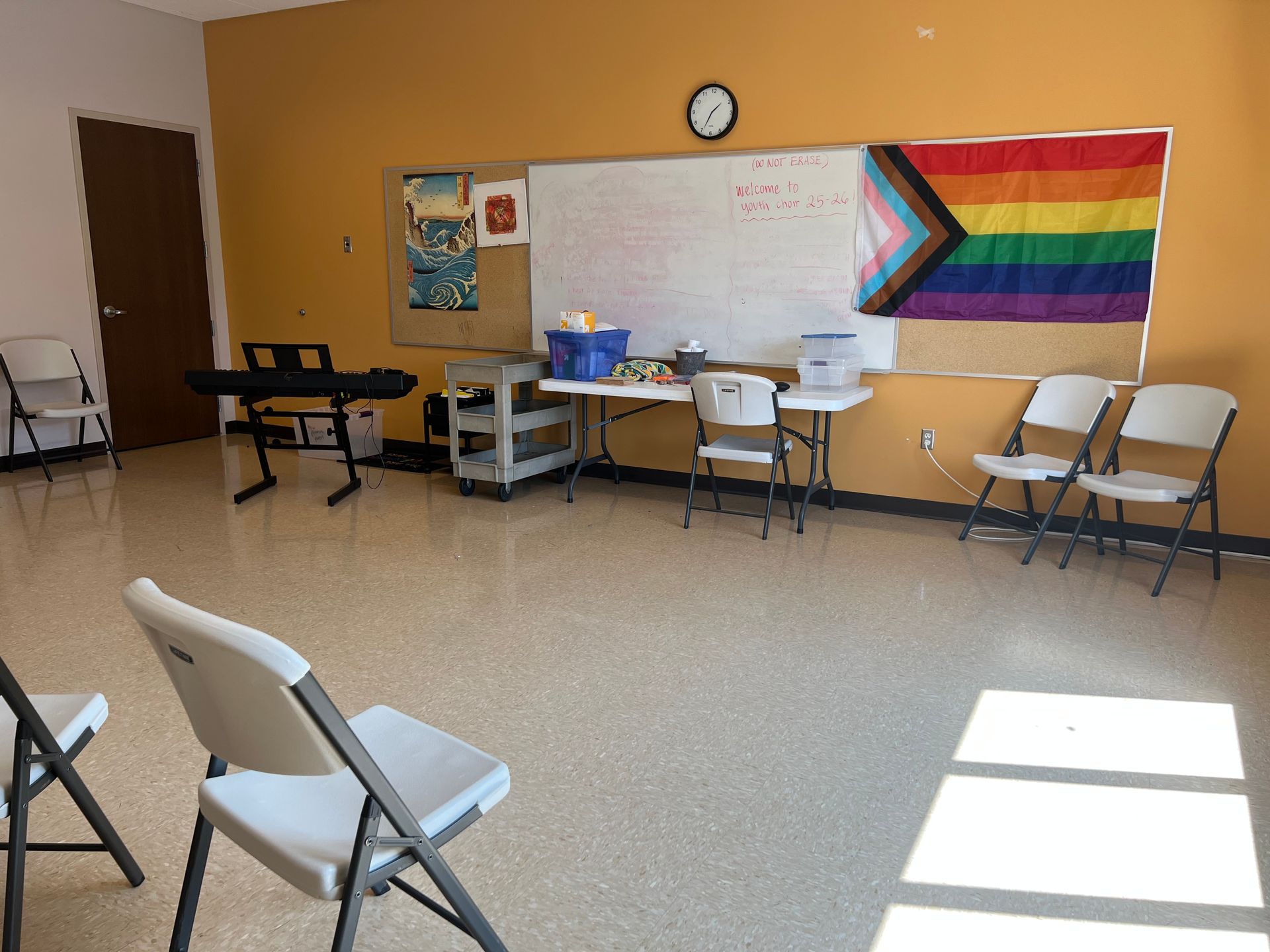 Community room with tables and folding chairs, wall clock, and rainbow Pride flag display.