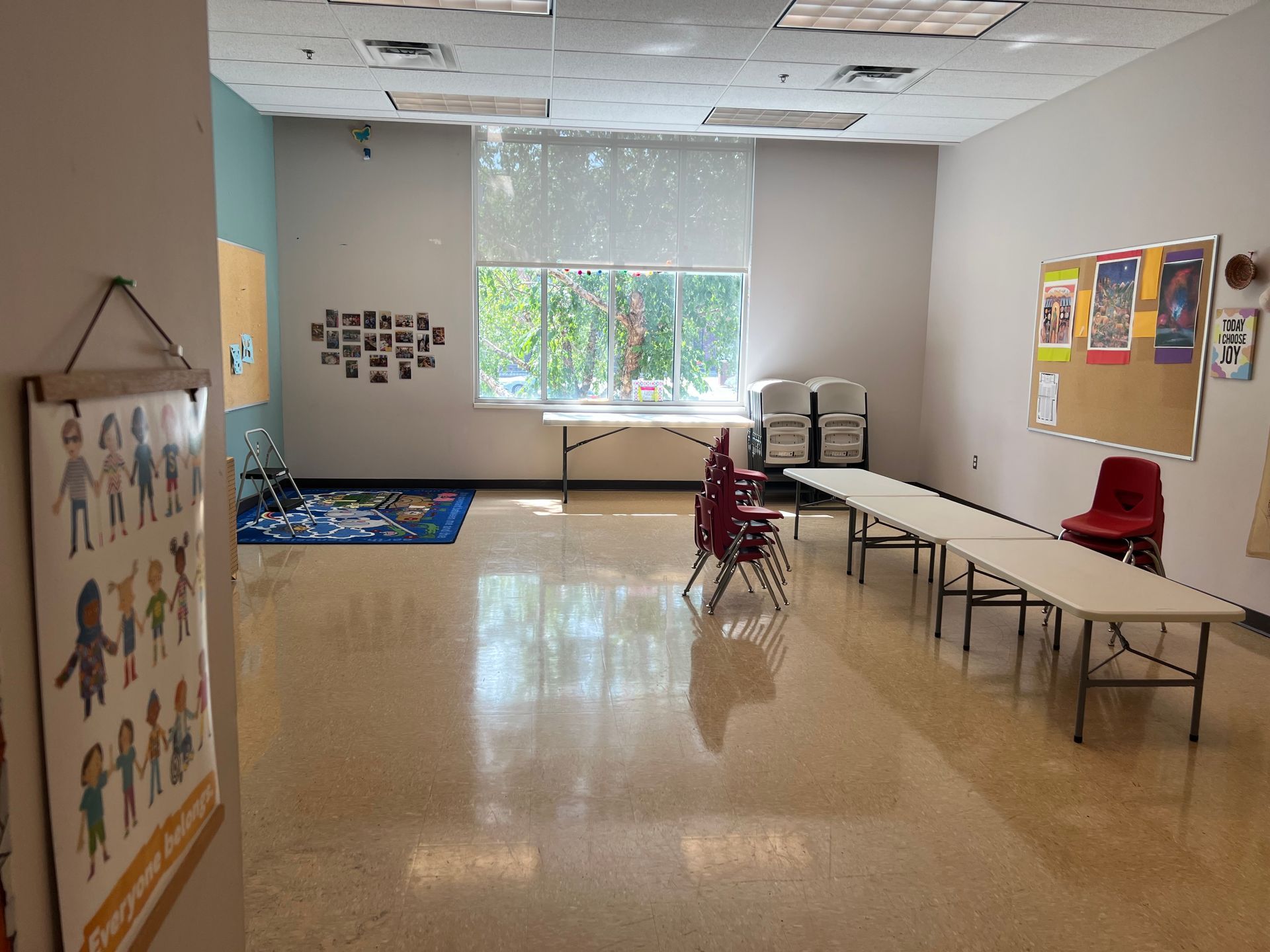 Empty classroom with tables, a red chair, colorful posters, and a windowed back wall.