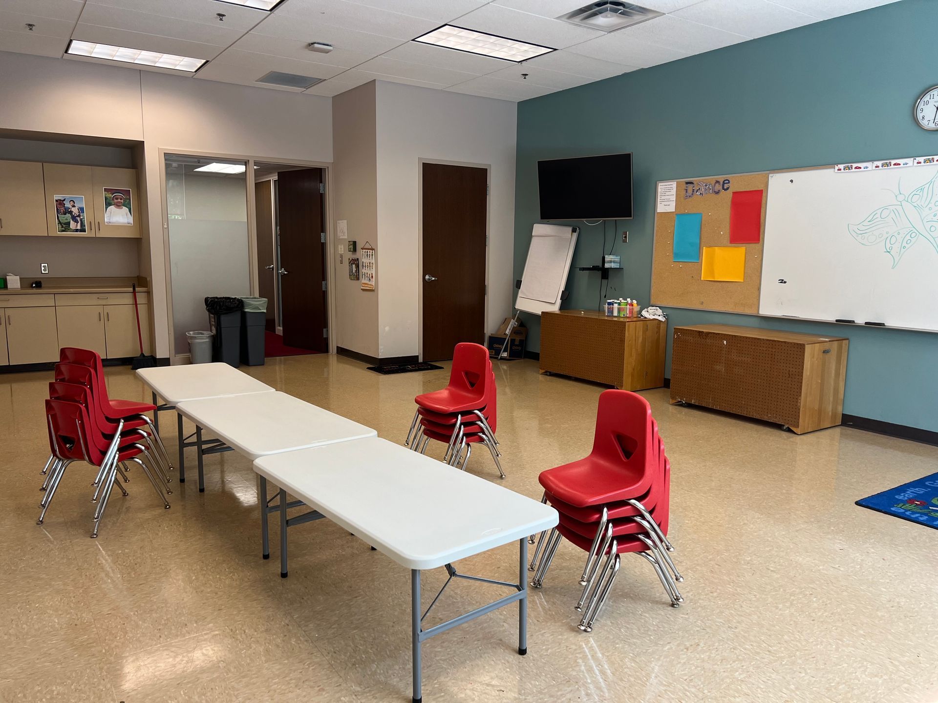 Empty classroom with red chairs around a white table, whiteboard, cabinets, and wall-mounted TV.