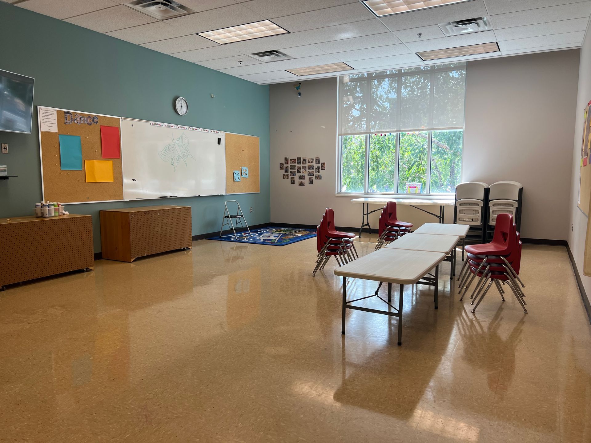 Empty classroom with tables and chairs, whiteboard, bulletin board, and large window letting in daylight