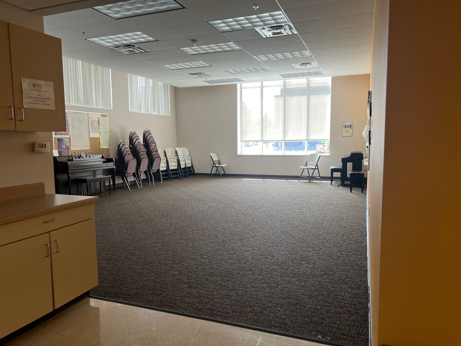Empty office room with chairs along the walls, carpeted floor, and bright windows at the back.