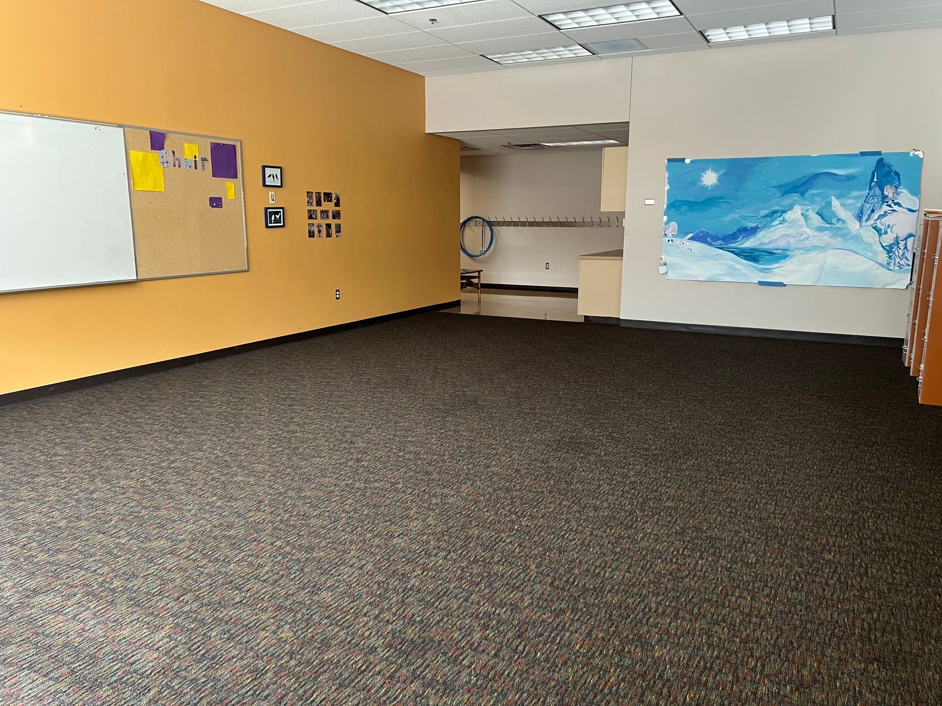 Empty classroom with tan walls, whiteboard, dark carpet, and a blue mural on the back wall