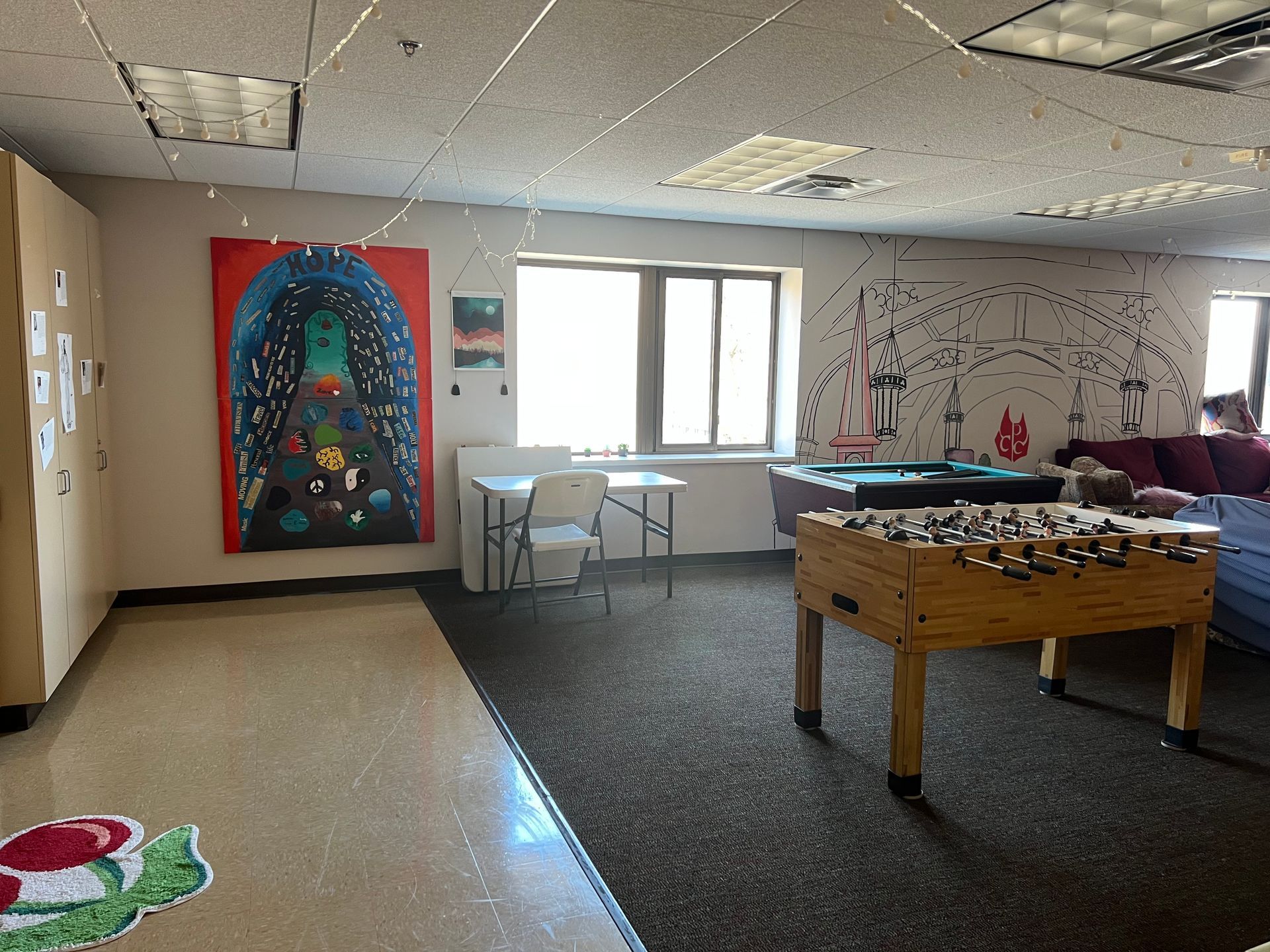 Community room with a foosball table, tables by a window, and colorful wall murals.