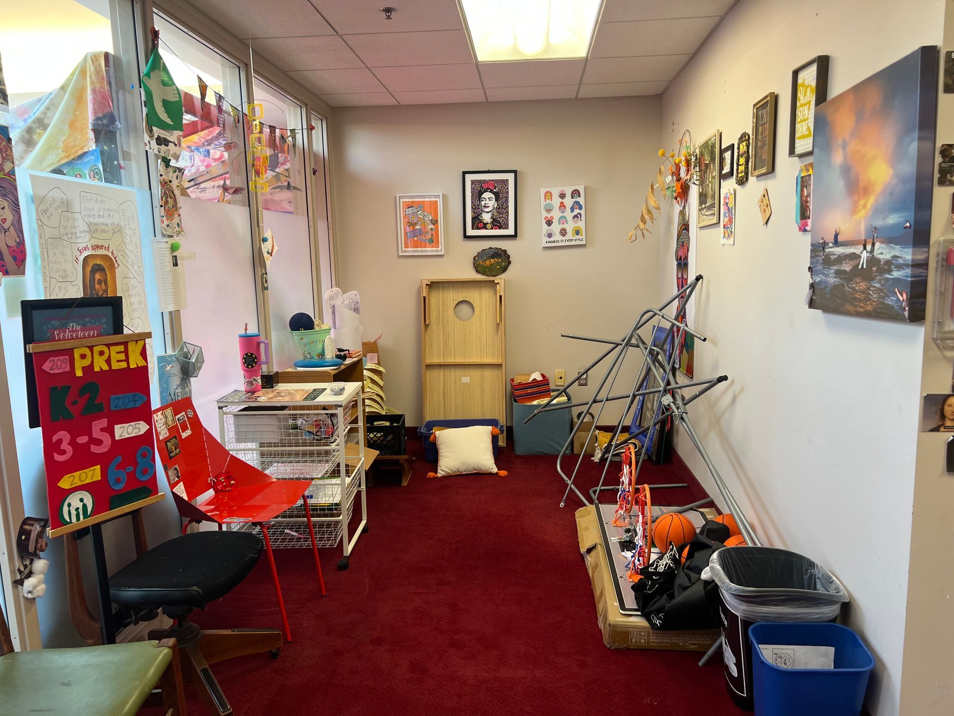 Narrow cluttered room with red carpet, bikes, shelves, wall art, and a PRE-K sign