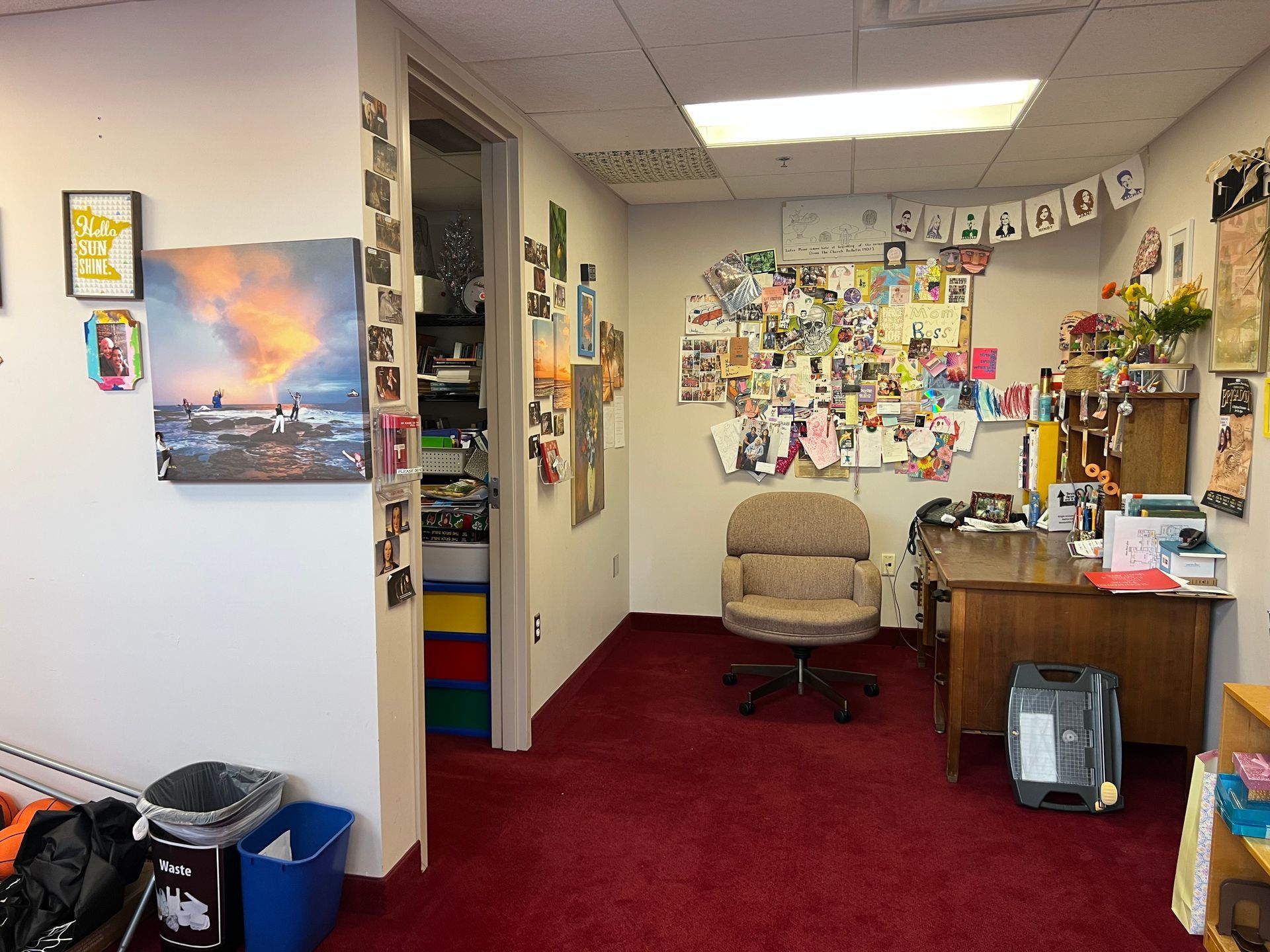 Office interior with burgundy carpet, desk, chair, wall art, and a bulletin board covered in notes and photos