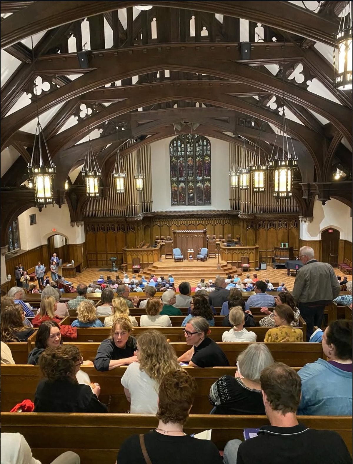 Church sanctuary with people seated in pews, facing a wood-paneled stage under arched beams.