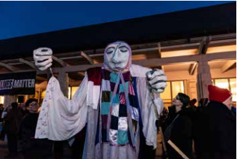 Person in a white ghost costume with colorful scarf stands outdoors at a nighttime event.