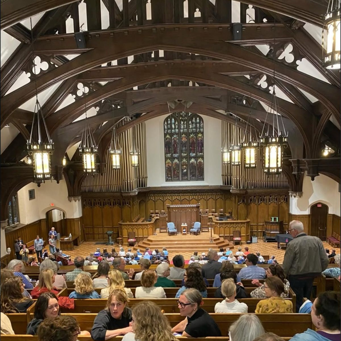 Wooden hall with arched beams and stained glass, filled with seated attendees at a gathering