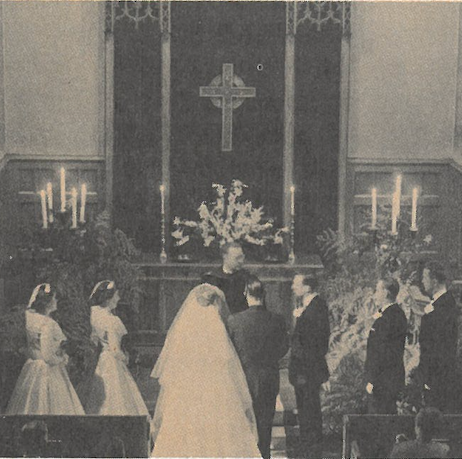 Wedding couple before altar in a church, with guests standing and a cross above the flowers