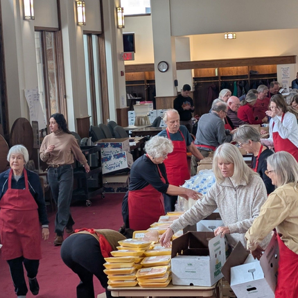 Volunteers handing out stacked food containers in a busy community hall