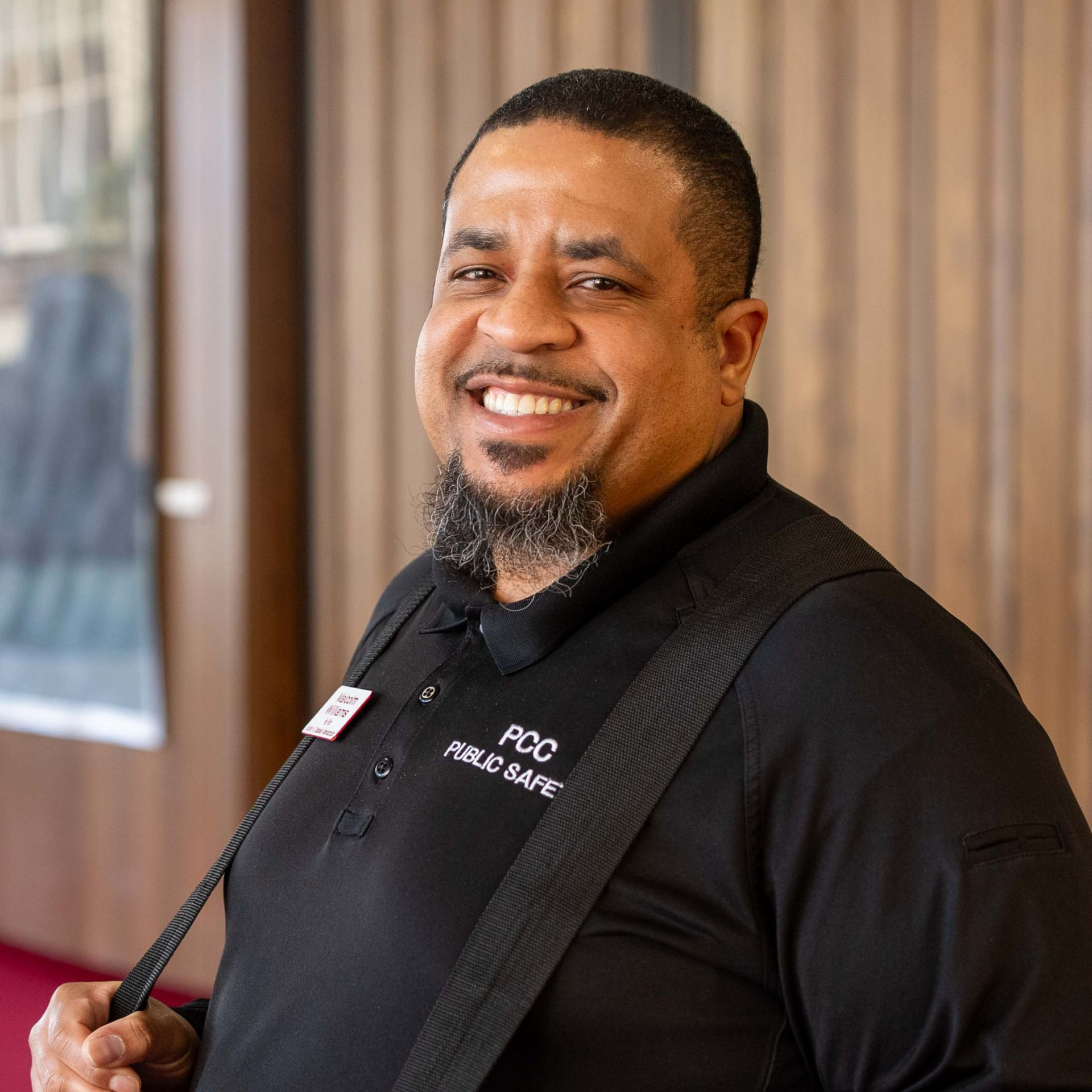 Smiling security officer in a black uniform standing indoors with a shoulder strap badge.