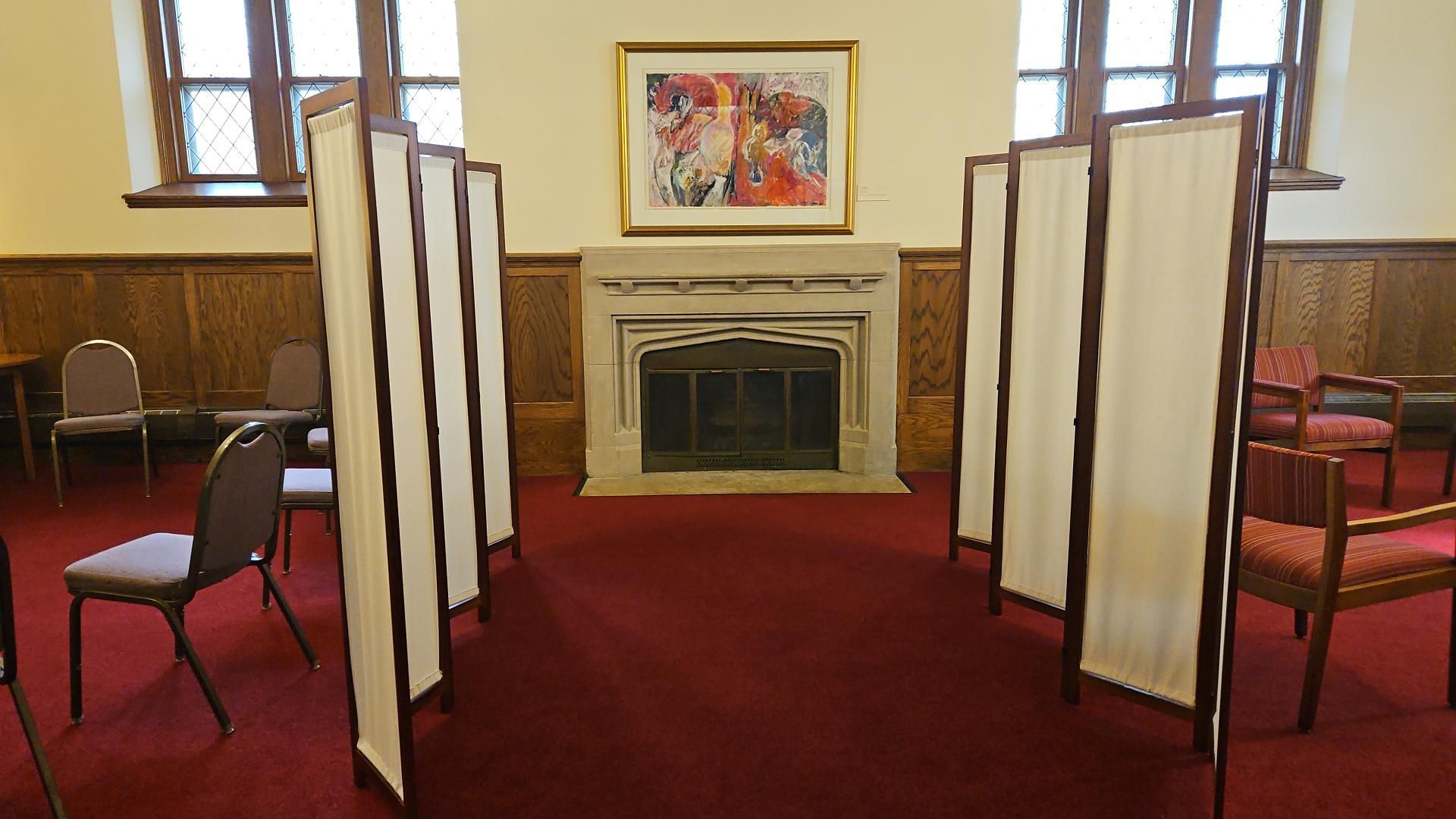 Red-carpeted room with folding privacy screens arranged around a fireplace and framed painting.