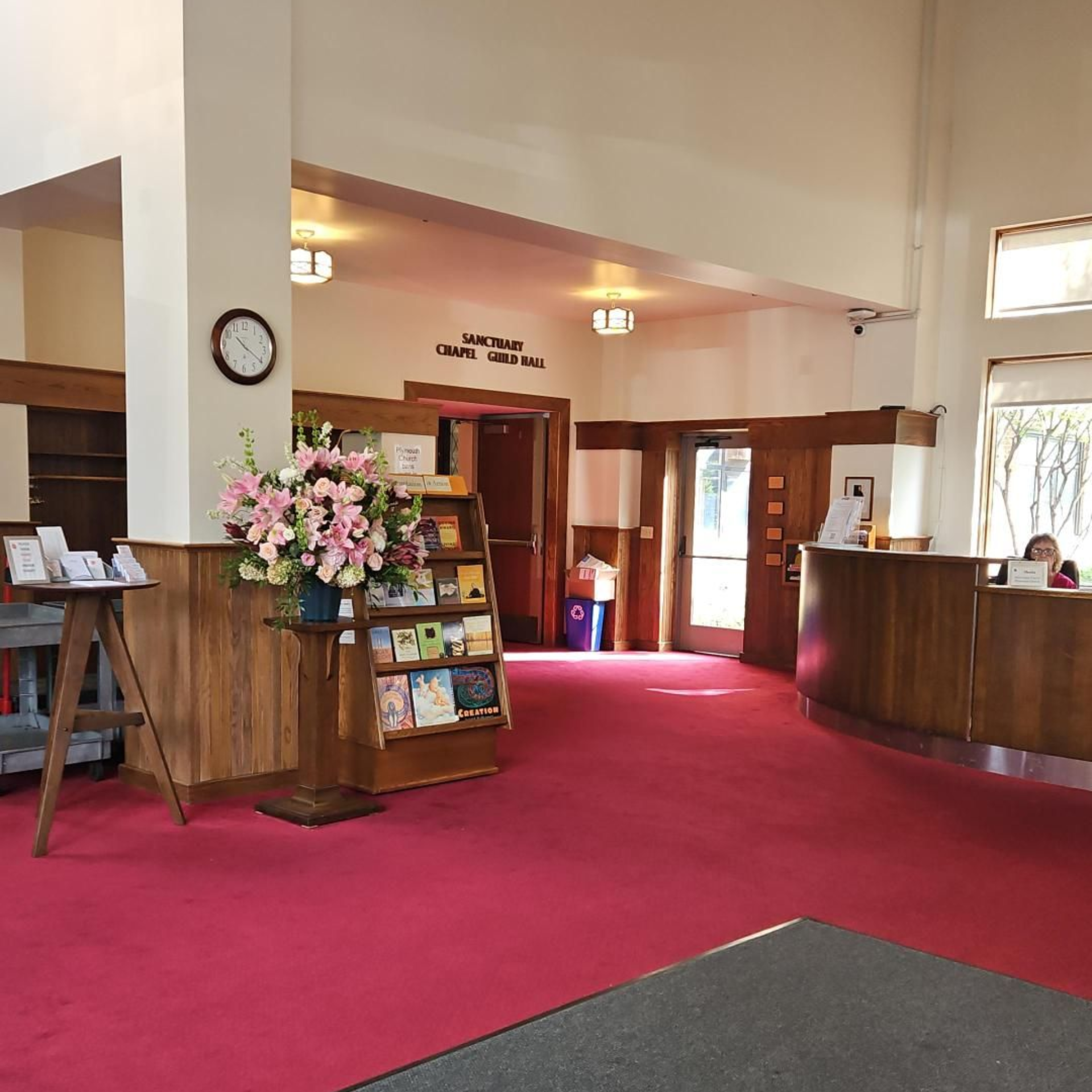 Brightly lit church sanctuary with rows of red chairs, wooden beams, and stained-glass windows