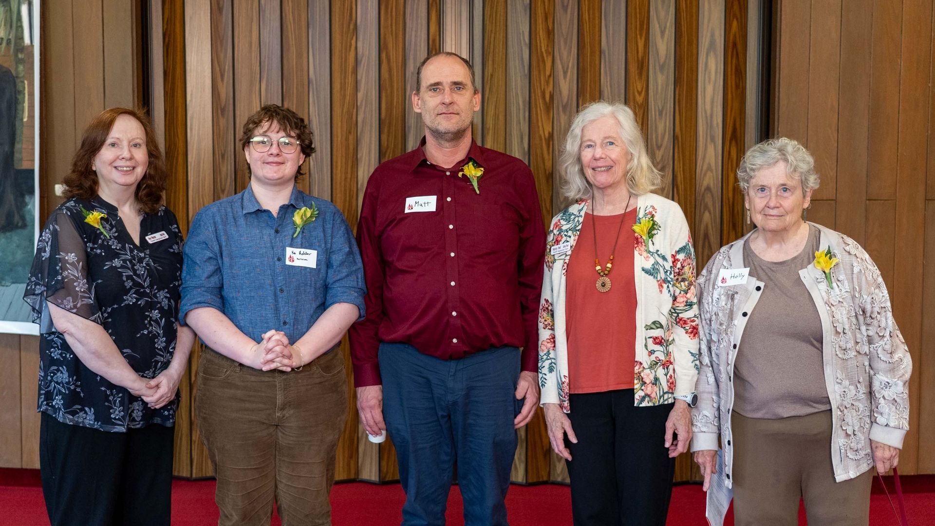 Five people standing indoors in front of a wooden wall, posing for a group photo.