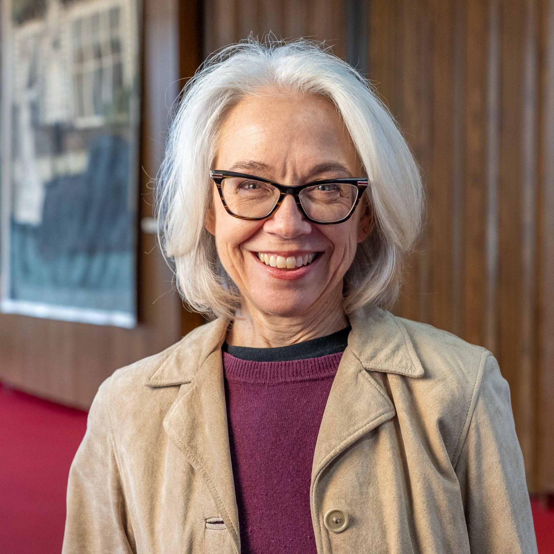Smiling woman with white hair and glasses standing by a wooden wall and red carpet