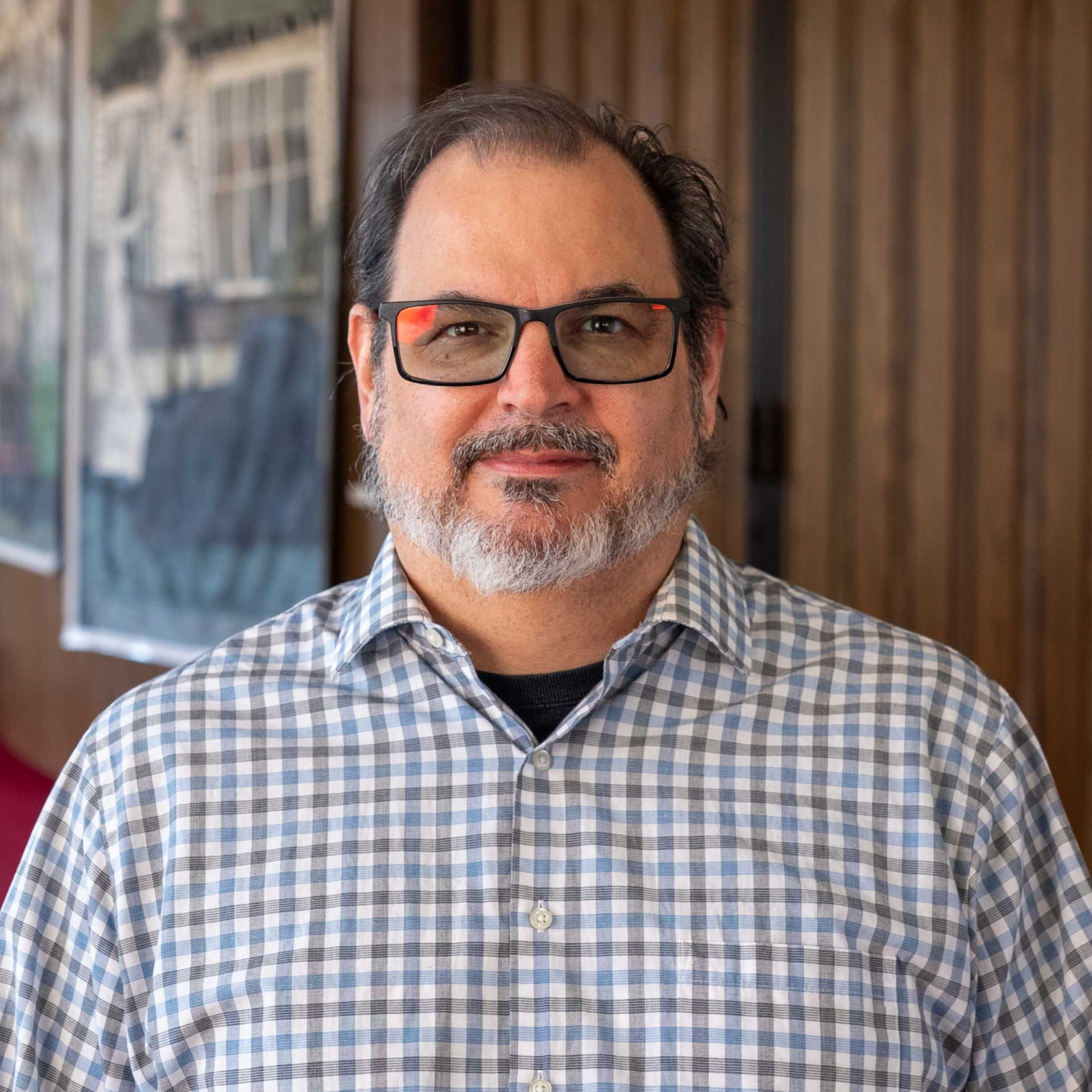 Man wearing glasses and a plaid shirt, smiling indoors against a blurred wall.