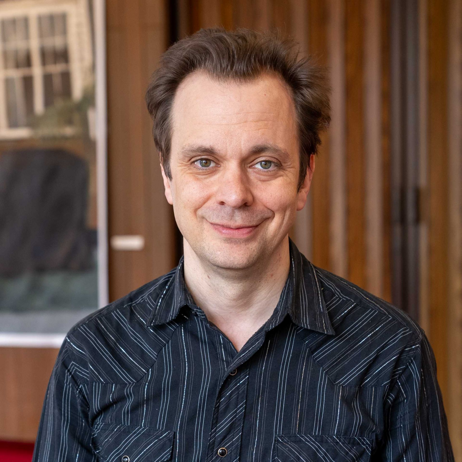 Man in a dark patterned shirt smiling in front of wooden paneling.