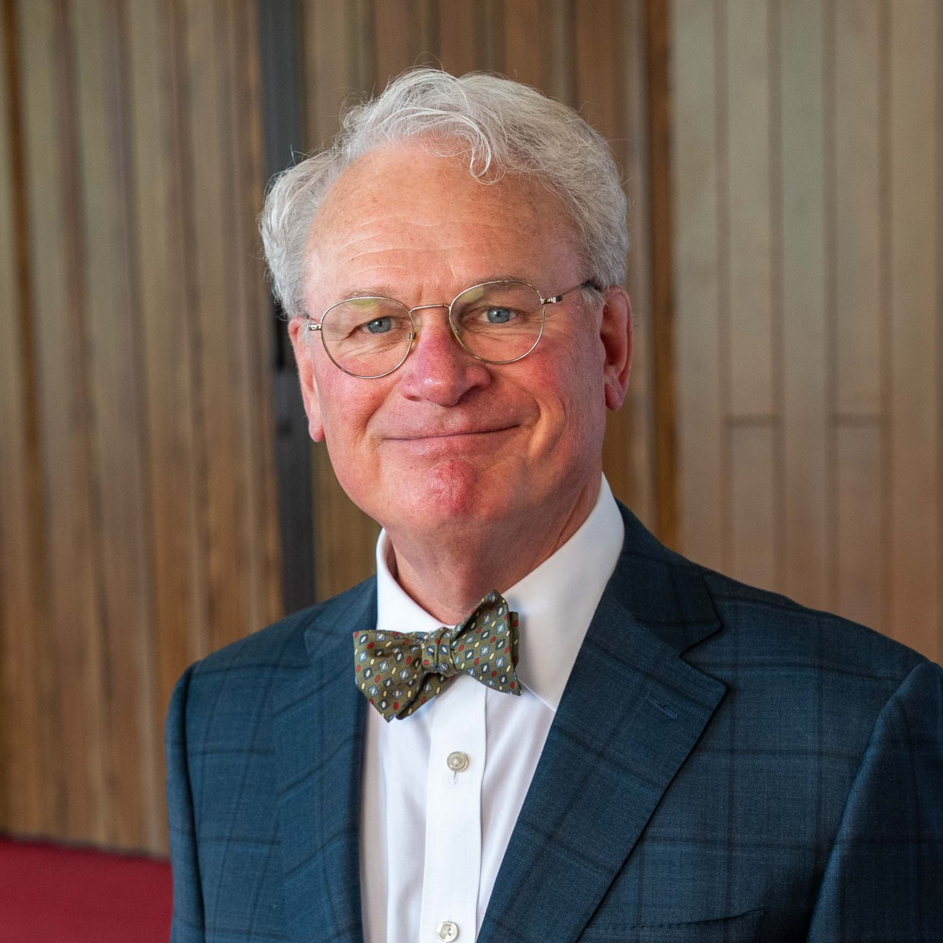 Portrait of a smiling man in glasses and a bow tie against a wood-paneled background